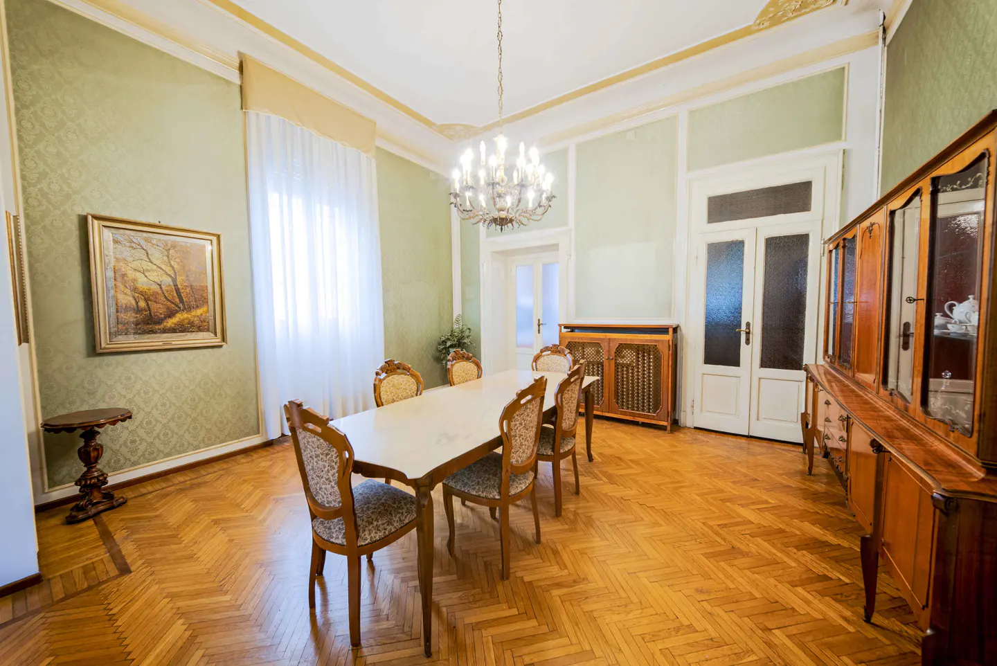 A dining room with parquet floors, light green walls, and a white table with six chairs. A chandelier hangs above the table. A painting and a wooden cabinet are also visible.