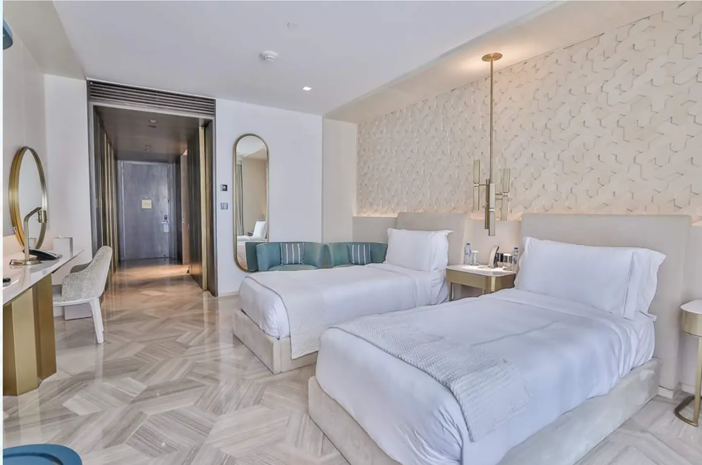 Hotel room with two twin beds, white linens, textured wall, gold accents, desk, and blue sofa.