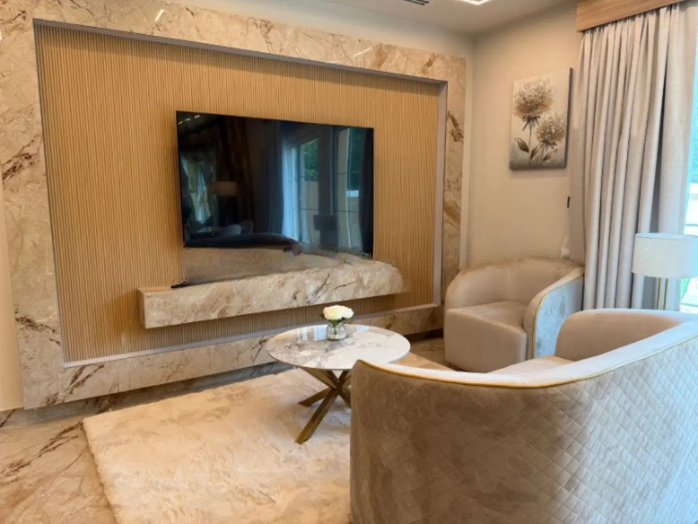 Living room with a TV mounted on a marble wall, two beige armchairs, and a marble coffee table with white roses.