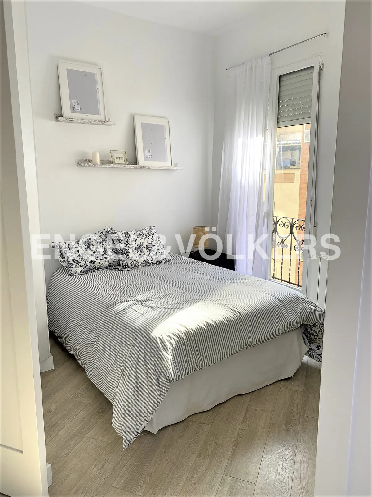 Bright bedroom with a striped duvet on the bed, white walls, and light wood floors. Shelves with picture frames are above the bed. Balcony with sheer curtains.