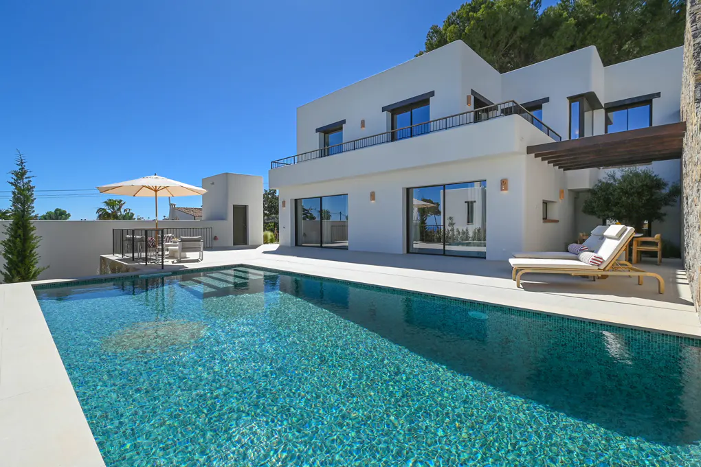 Modern white villa with a turquoise pool under a blue sky. Lounge chairs and an umbrella offer relaxation.