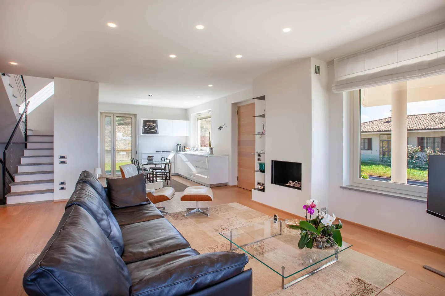 Bright, open-concept living room with a black leather sofa, glass coffee table, and a view of the kitchen and staircase. A fireplace and large window add to the modern design.