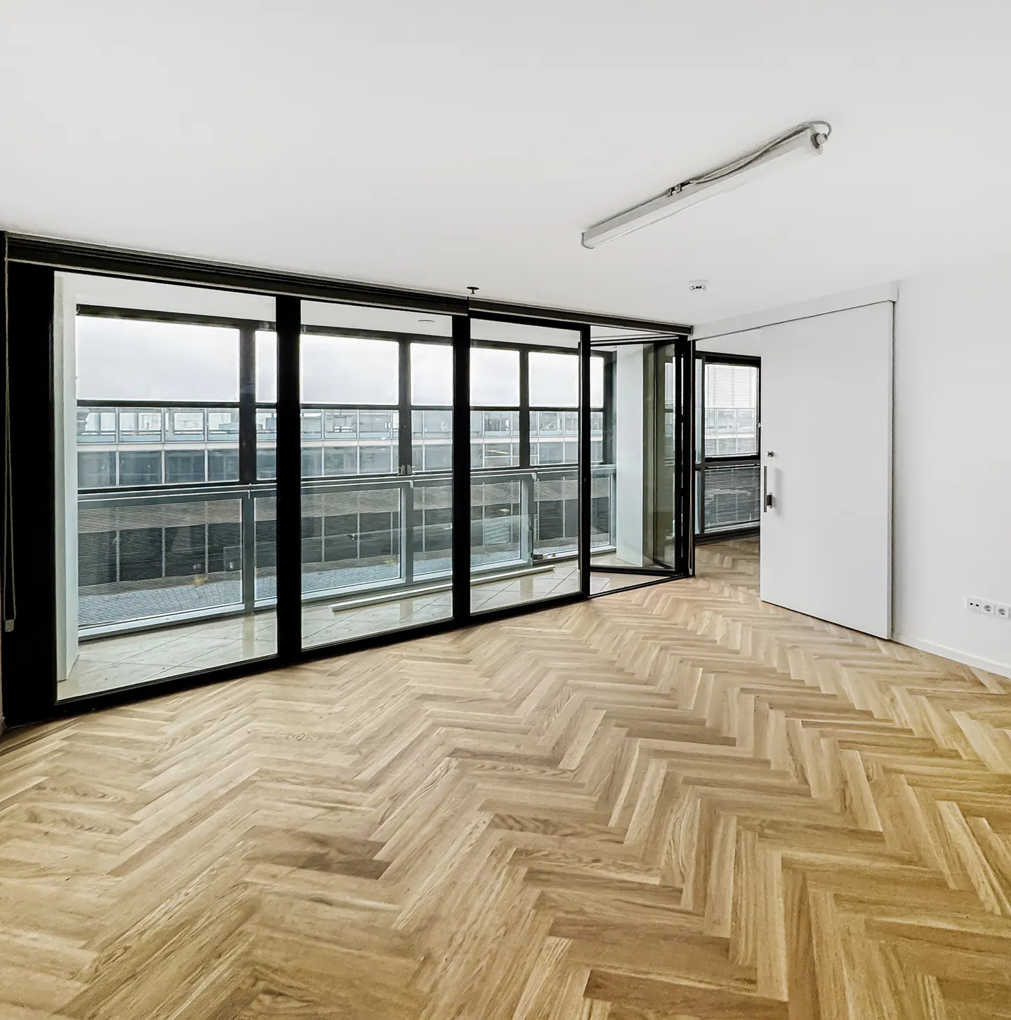 An empty room with herringbone wood floors, white walls, and a large window with a view of a building.