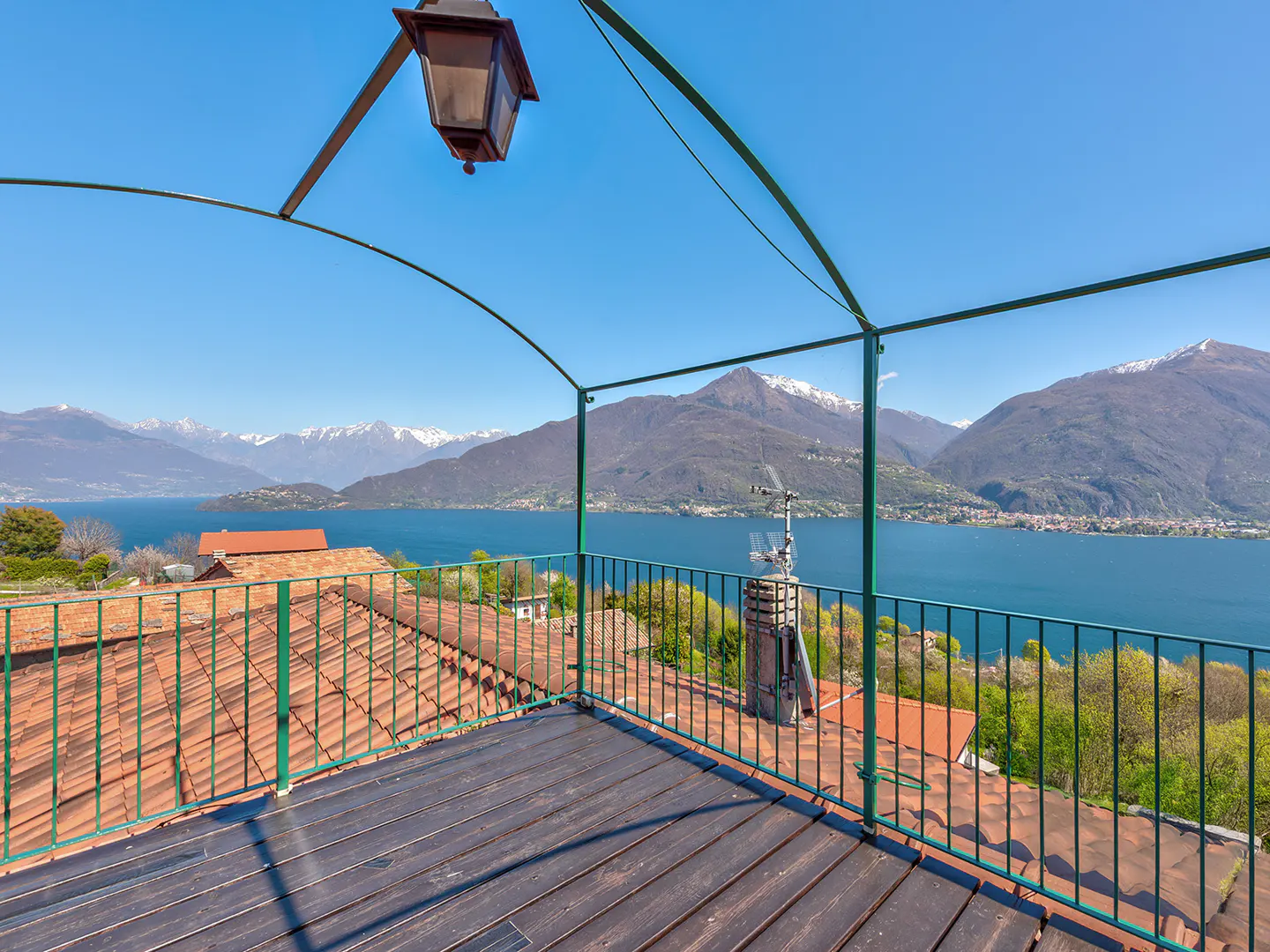 A balcony with a green railing overlooks a lake and mountains under a blue sky. A vintage lamp hangs above the wooden floor.