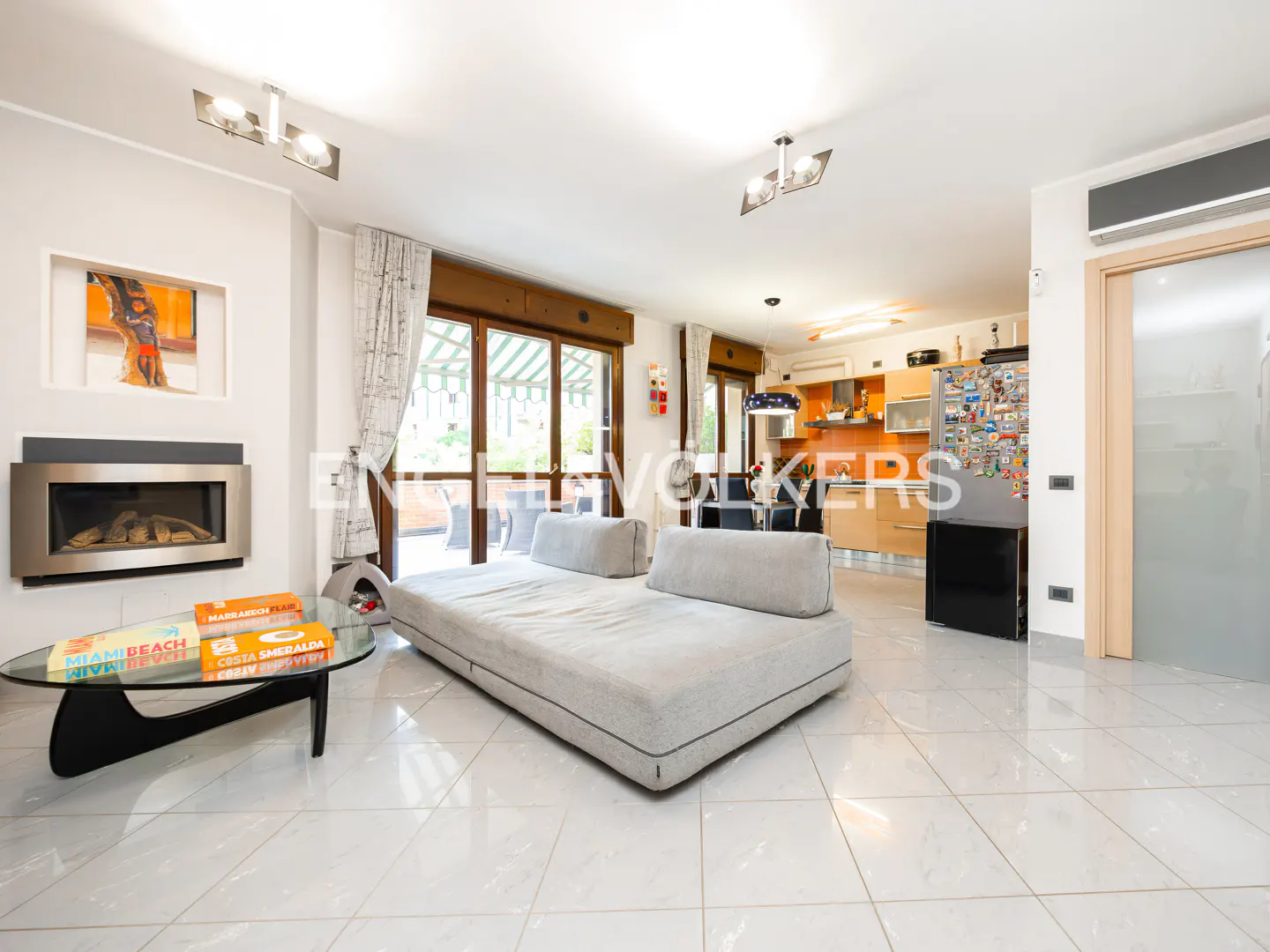 A bright living room with a gray sofa, fireplace, and a modern glass coffee table. The kitchen is visible in the background.