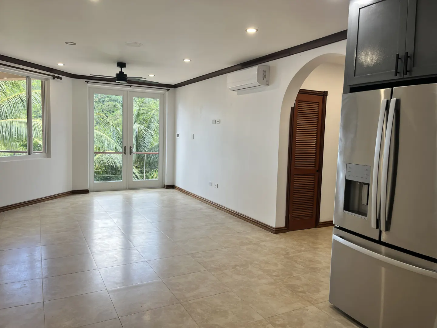 Bright, empty living room with tile floors, white walls, and a stainless steel refrigerator. Balcony doors open to lush greenery.