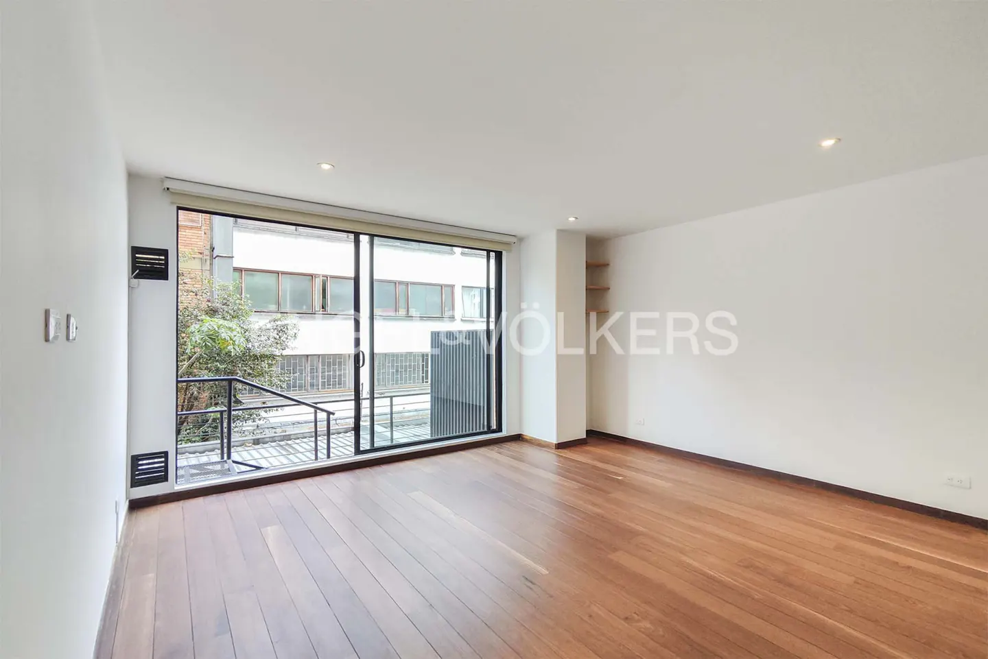 Empty room with white walls, polished wood floors, and a large sliding glass door looking out onto a balcony and building.