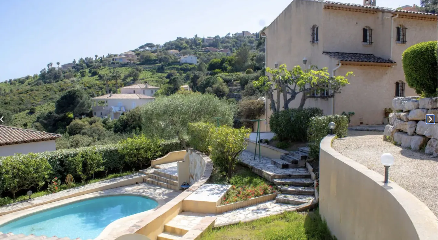 Exterior view of a tan house with a blue pool, stone steps, and green hillside with other houses in the background.