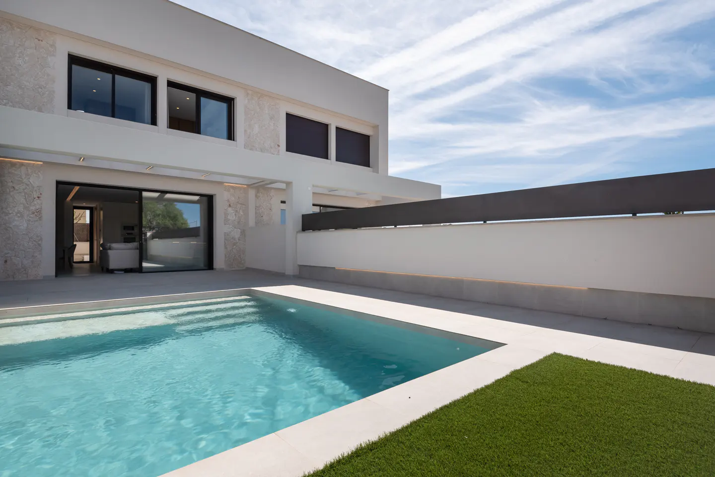 Modern white house with a turquoise pool, gray patio, and small patch of green grass under a blue sky with white clouds.