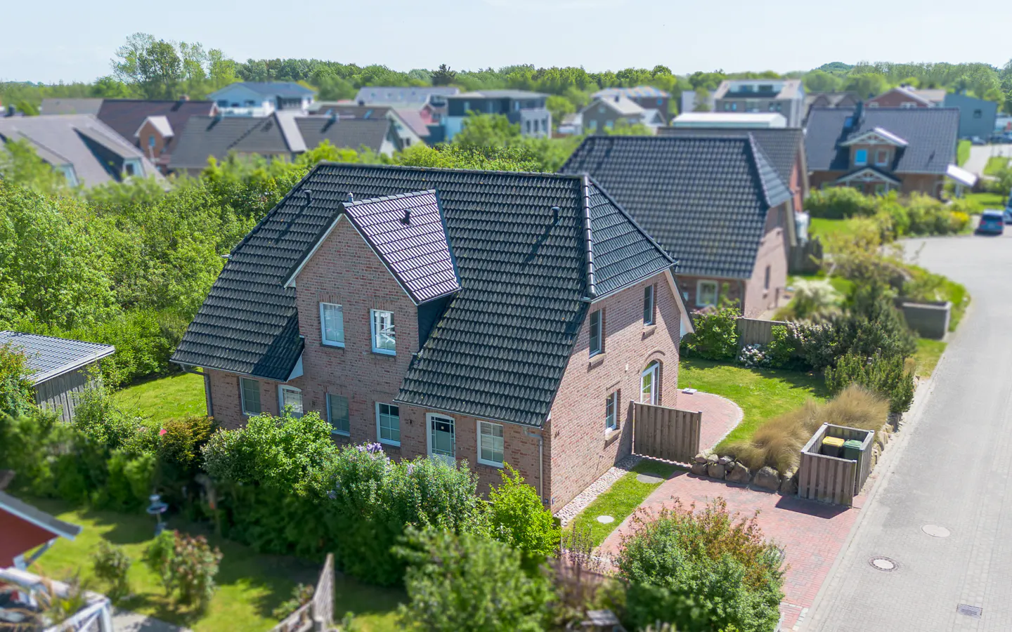Aerial view of a two-story brick house with a dark gray roof, surrounded by green trees and other houses in the background.