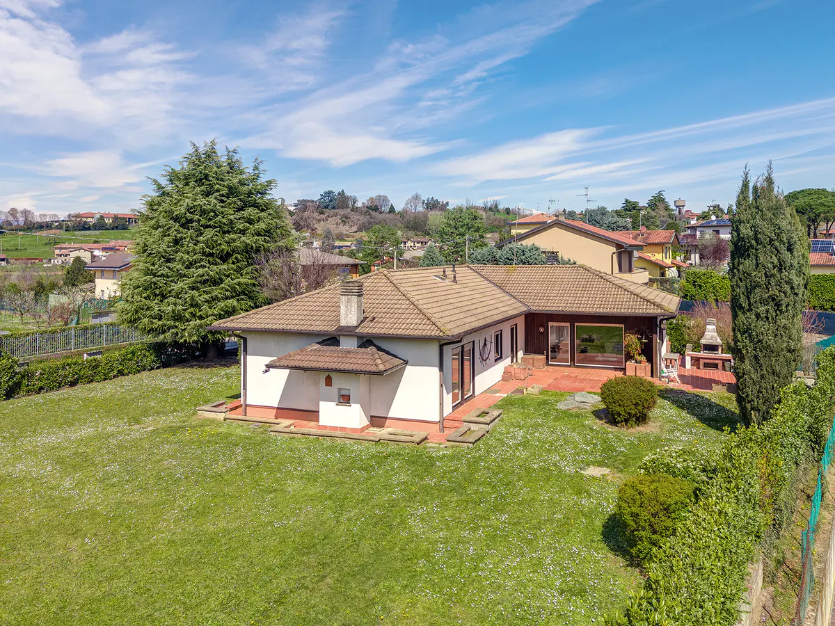 Aerial view of a one-story white house with a brown tile roof, a green lawn, and trees under a blue sky.