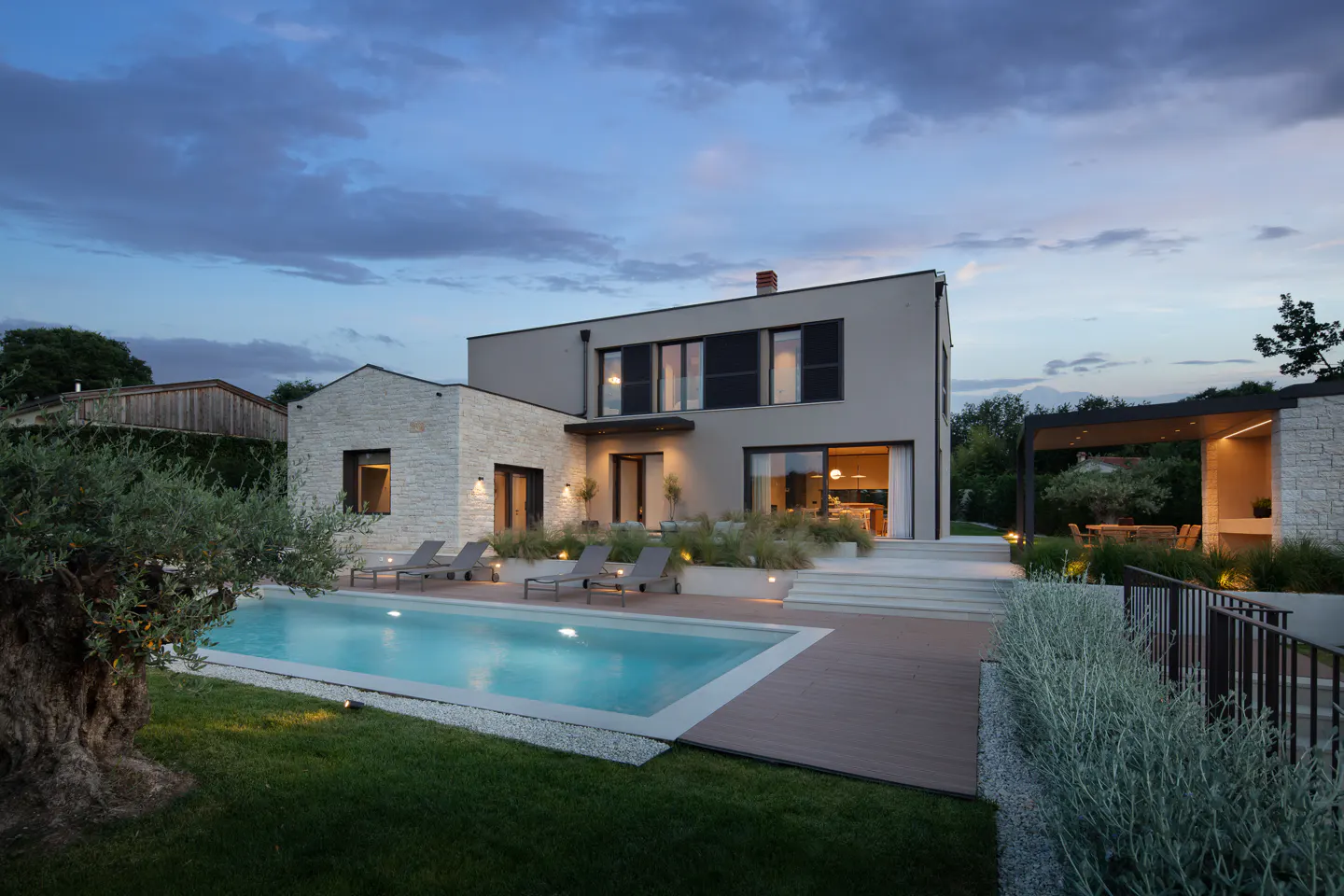 Modern two-story house with a pool, lounge chairs, and a patio at dusk. The house is beige and stone, with black window frames.