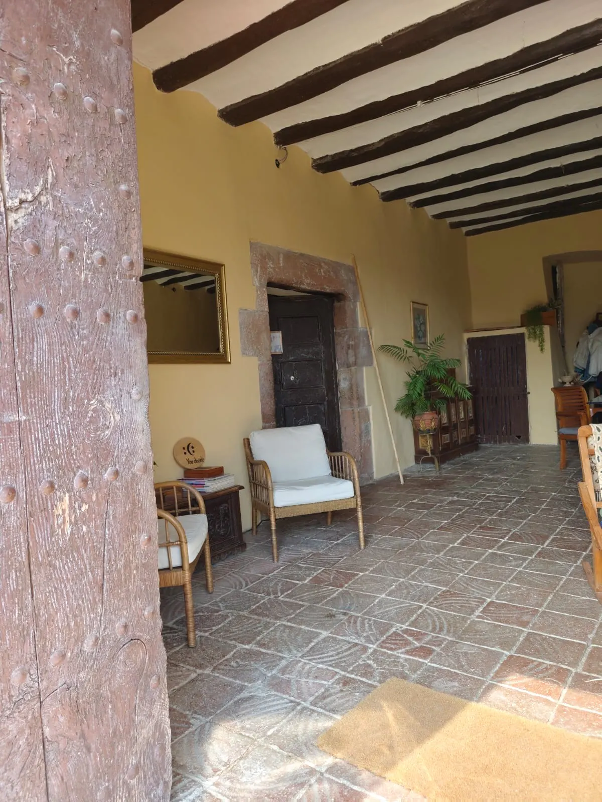 Covered patio with brown beams, tan walls, and brick floors. Wicker chairs with white cushions sit near a dark wood door.