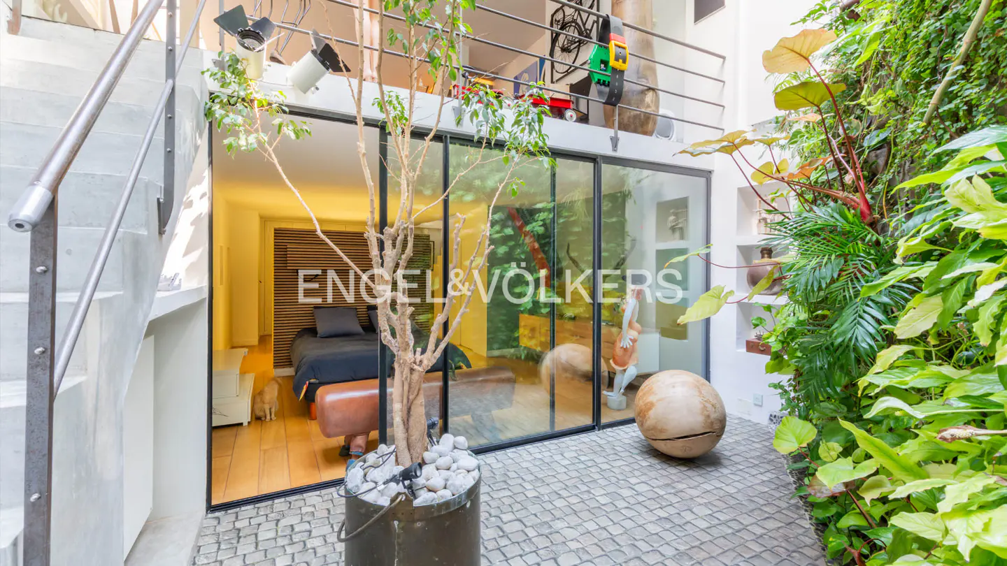 Indoor courtyard with stone pavers, lush greenery, and a glass-walled room. A tree in a pot sits near the glass, with stairs on the left.