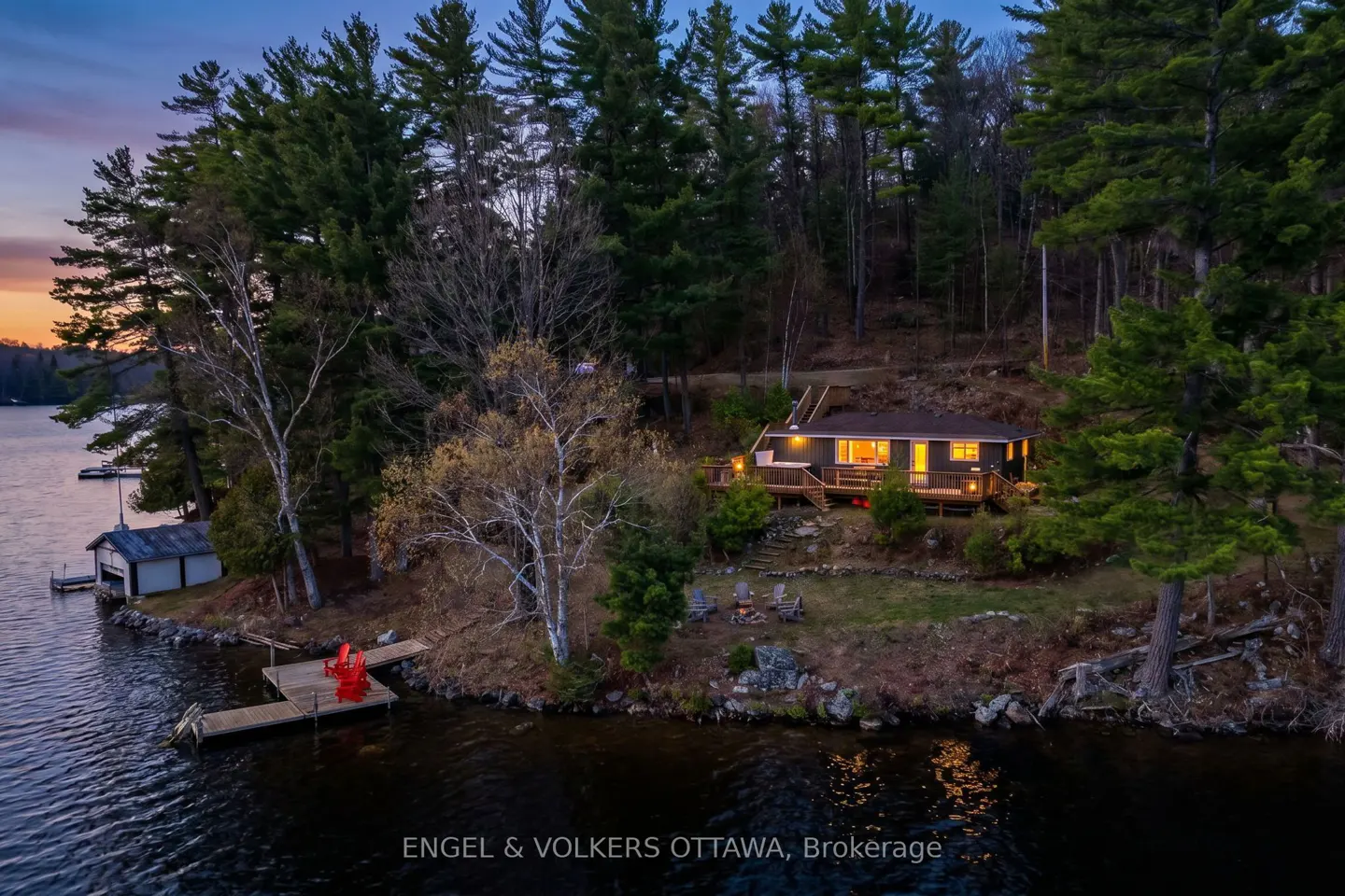 Aerial view of a lake house with a dock, red chairs, and a boathouse surrounded by trees at dusk.