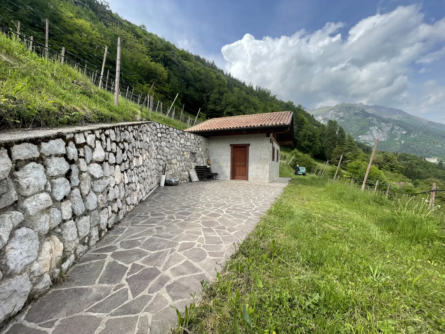 Stone cottage with a red tile roof, stone wall, and a mountain backdrop. A stone path leads to the cottage.