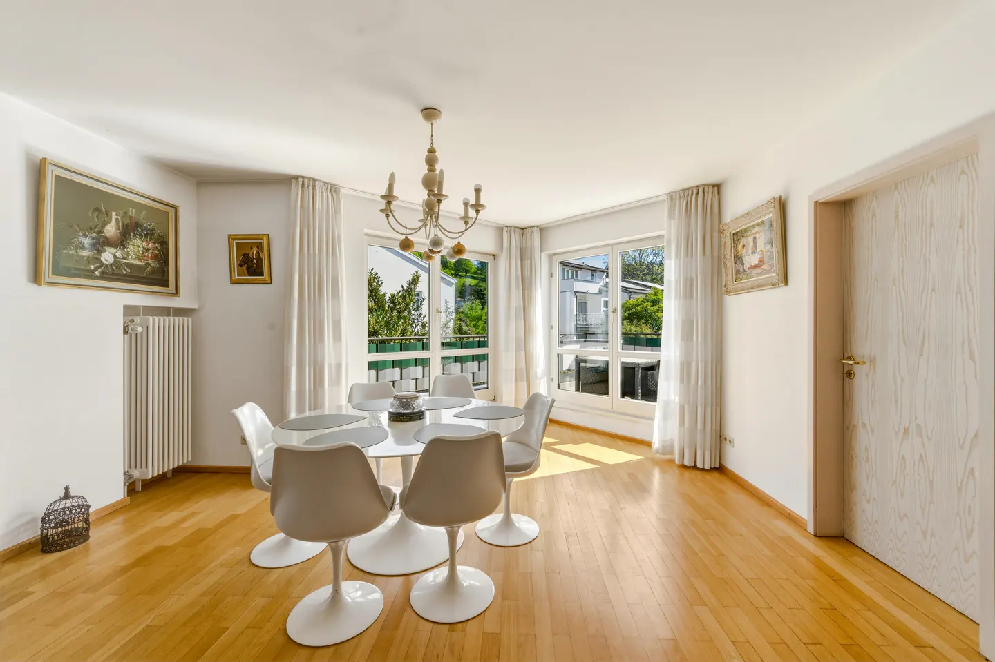 Bright dining room with wood floors, white walls, and a round white table with six chairs. Windows with white curtains overlook greenery.