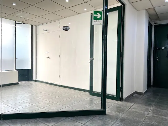 Empty office space with gray tile floor, white walls, and dark green door frames. An exit sign is visible above a doorway.