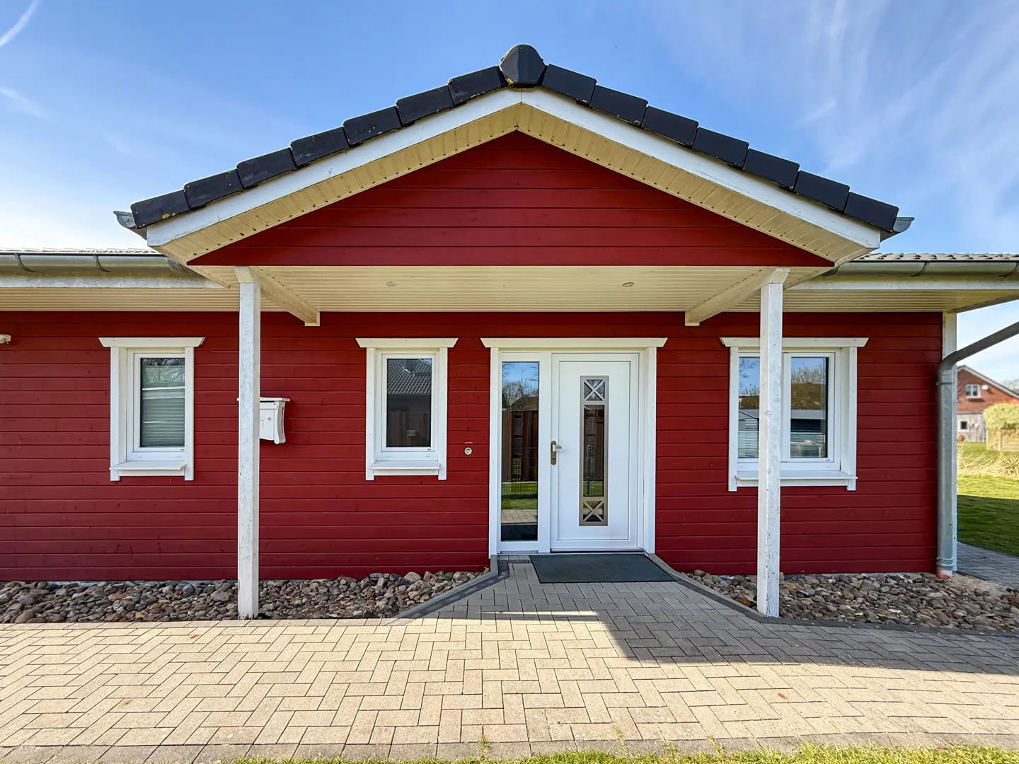 Red wooden house with white trim, a black roof, and a white front door. A brick walkway leads to the entrance.