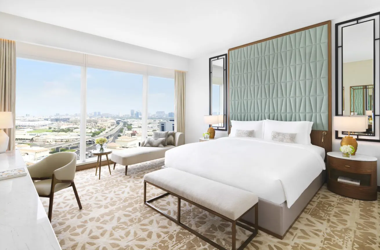 Modern hotel bedroom with a white bed, sage green tufted headboard, and floor-to-ceiling windows overlooking a city skyline.