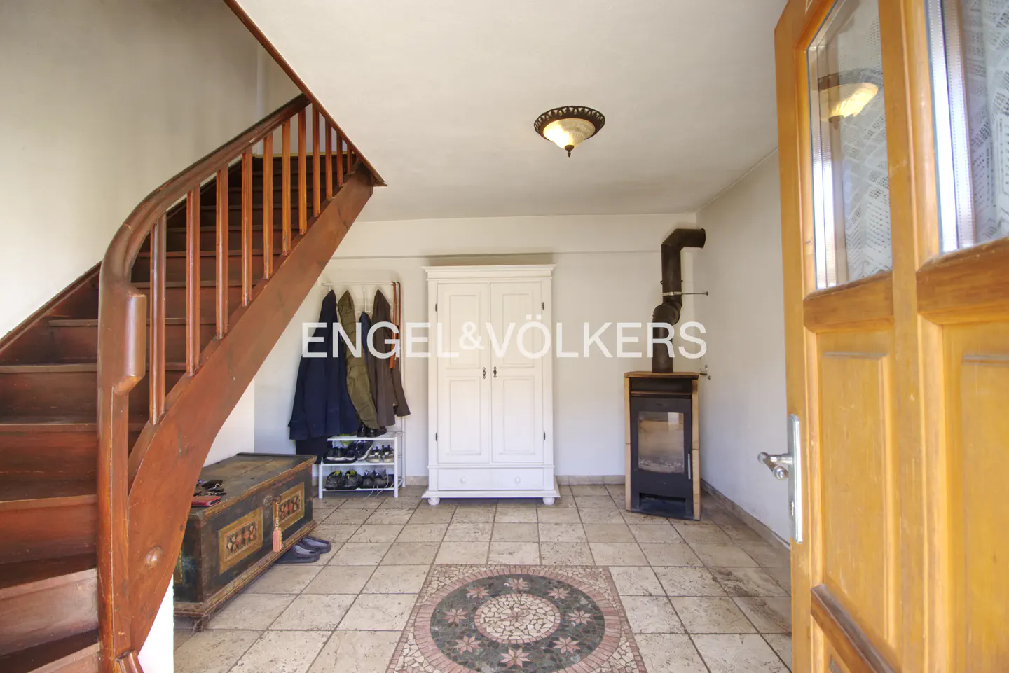 Entryway with wood staircase, tile floor, white wardrobe, wood-burning stove, and open wood door. Coats hang on a rack.
