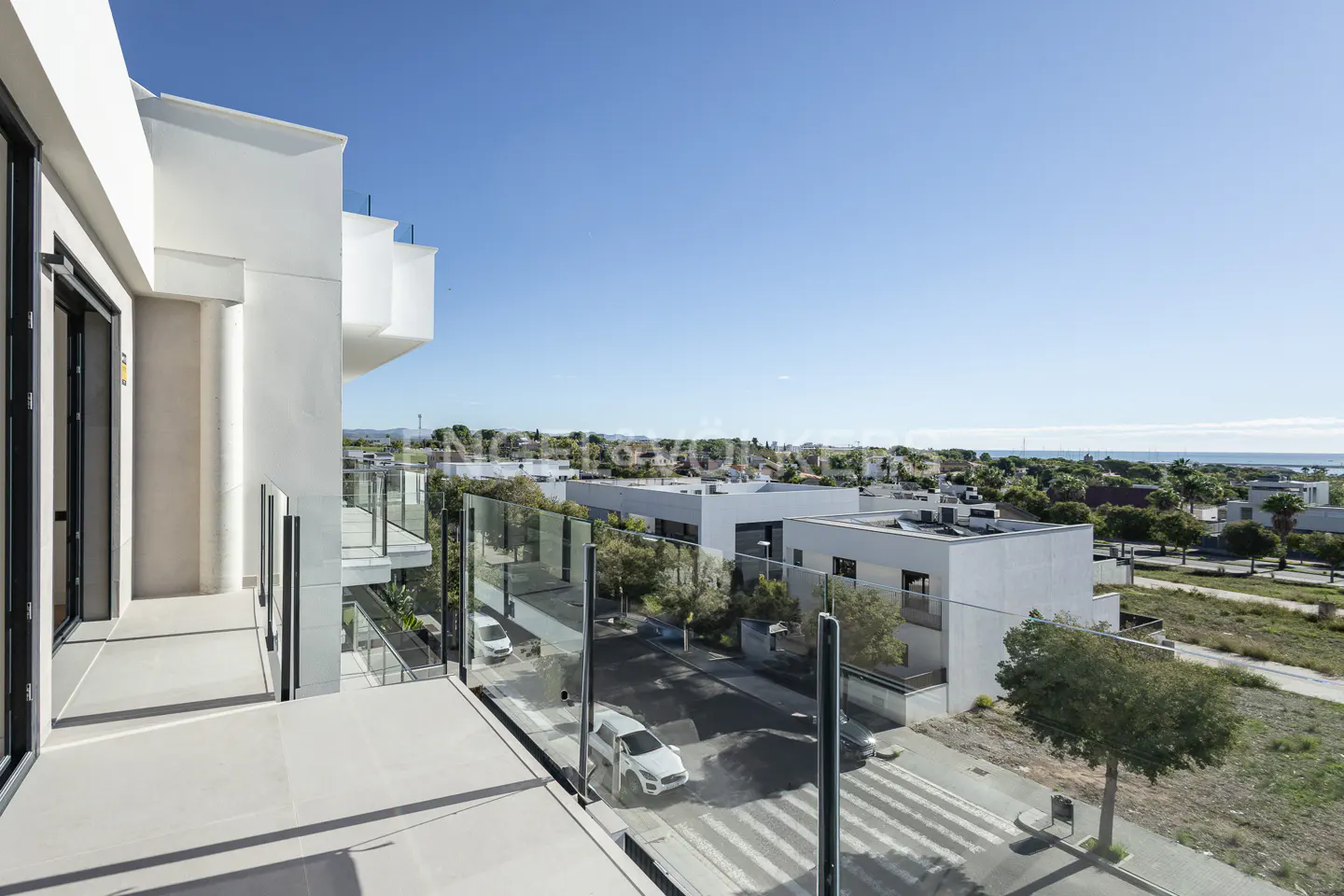 View from a modern balcony with glass railings overlooking a street with white buildings and a glimpse of the ocean.