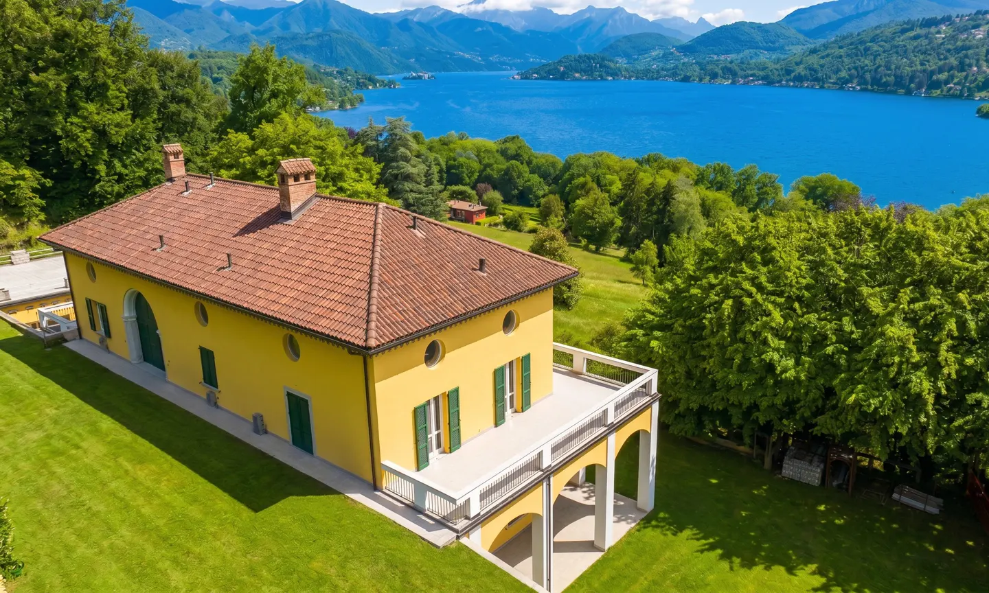 Aerial view of a yellow villa with a red tile roof, green shutters, and a lake and mountains in the background.