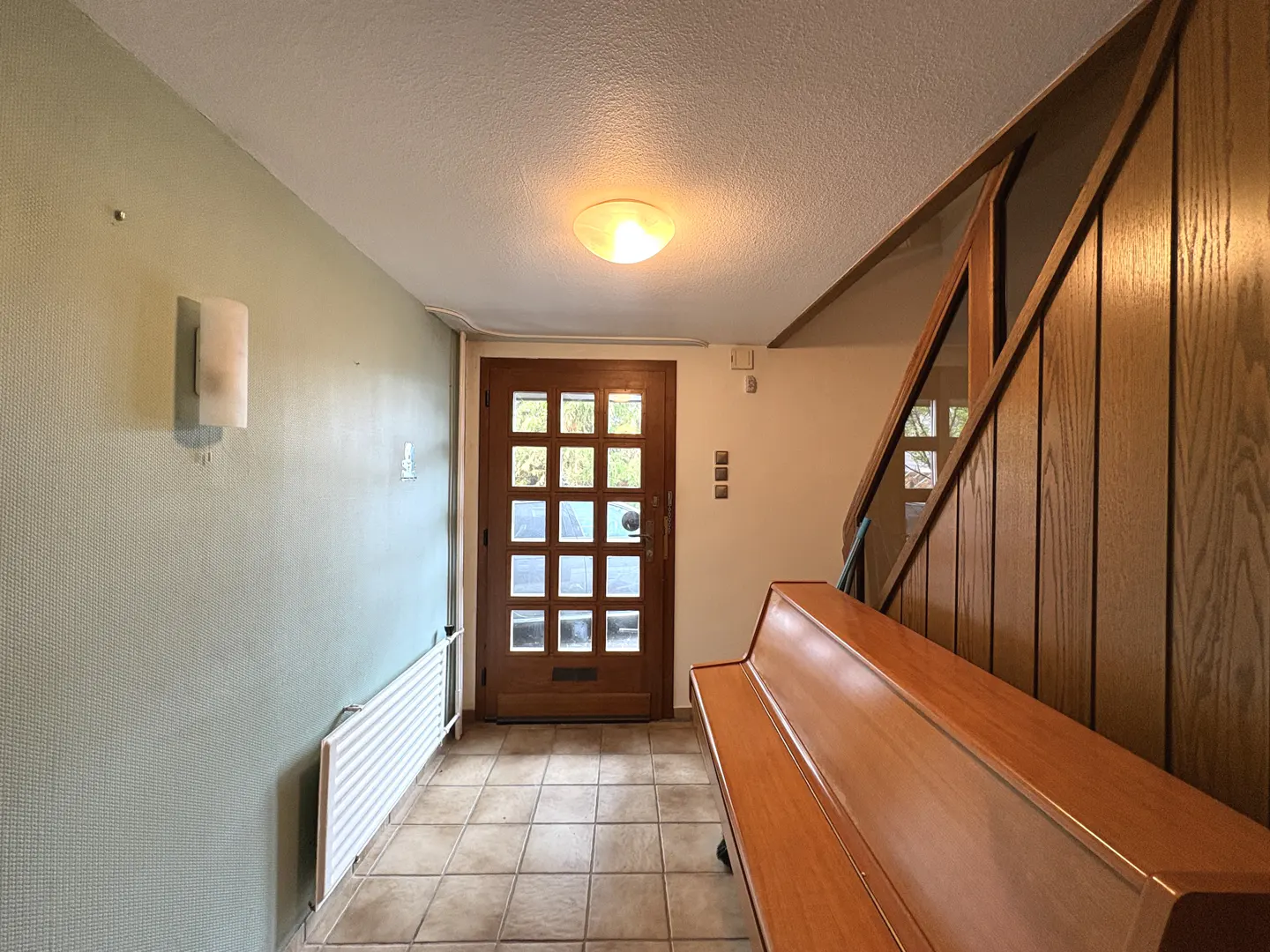 Entryway with a wood door, tiled floor, and a light green wall. A wood staircase and a bench are on the right.