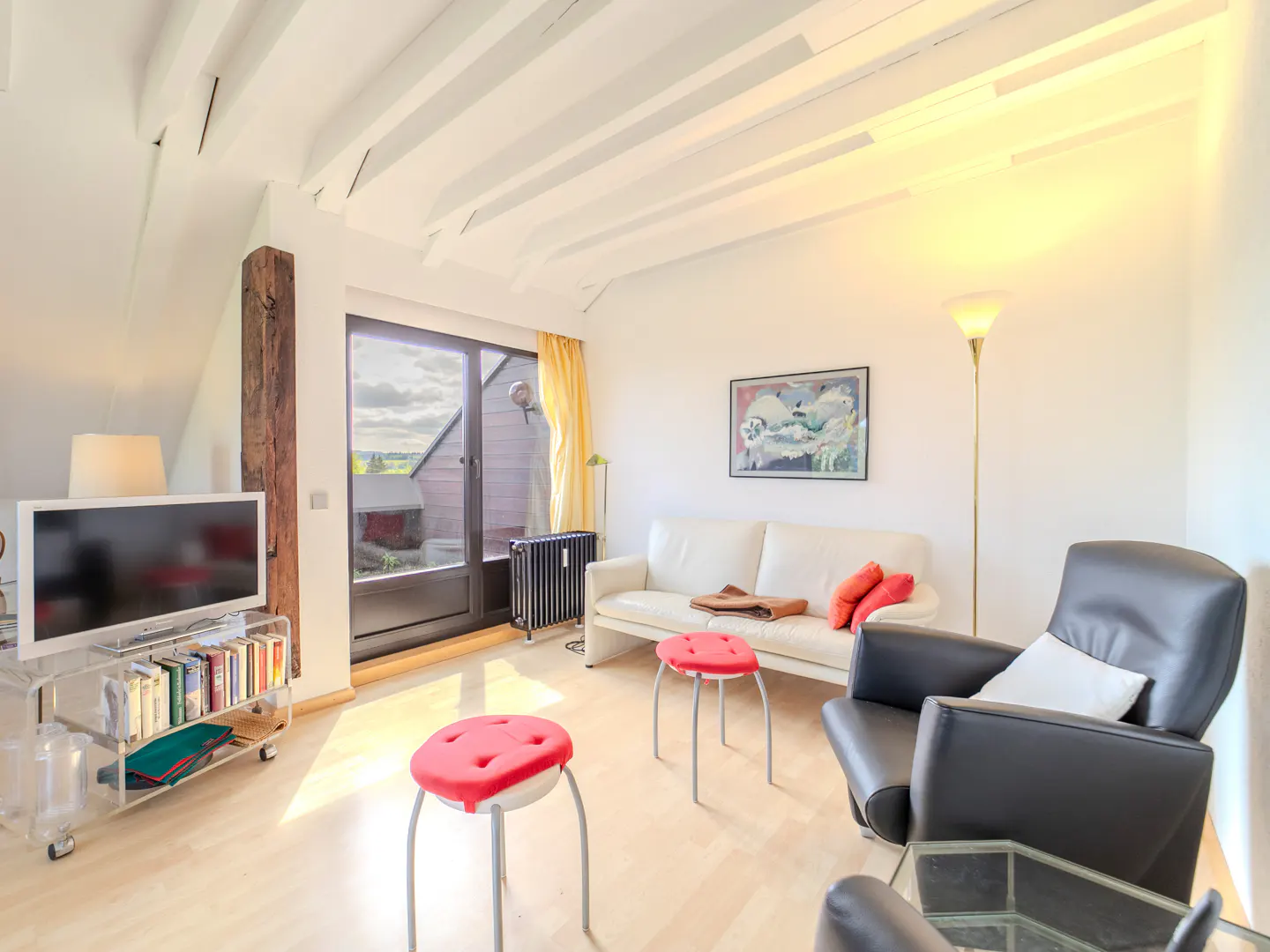 Bright living room with white walls, wood floors, and beamed ceiling. Features a white sofa, black chairs, and red stools. A TV sits on a clear stand.