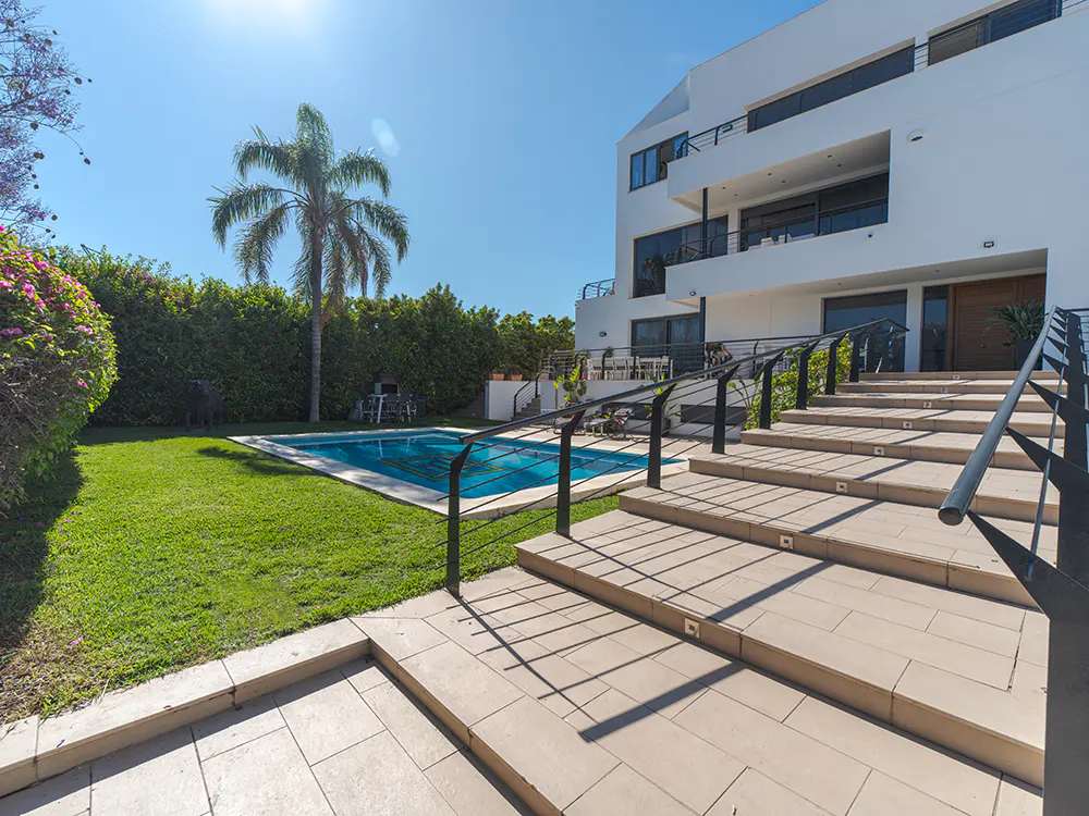 Modern white house with a pool. Stone steps with black railings lead up to the house. Palm tree and green lawn in the background.