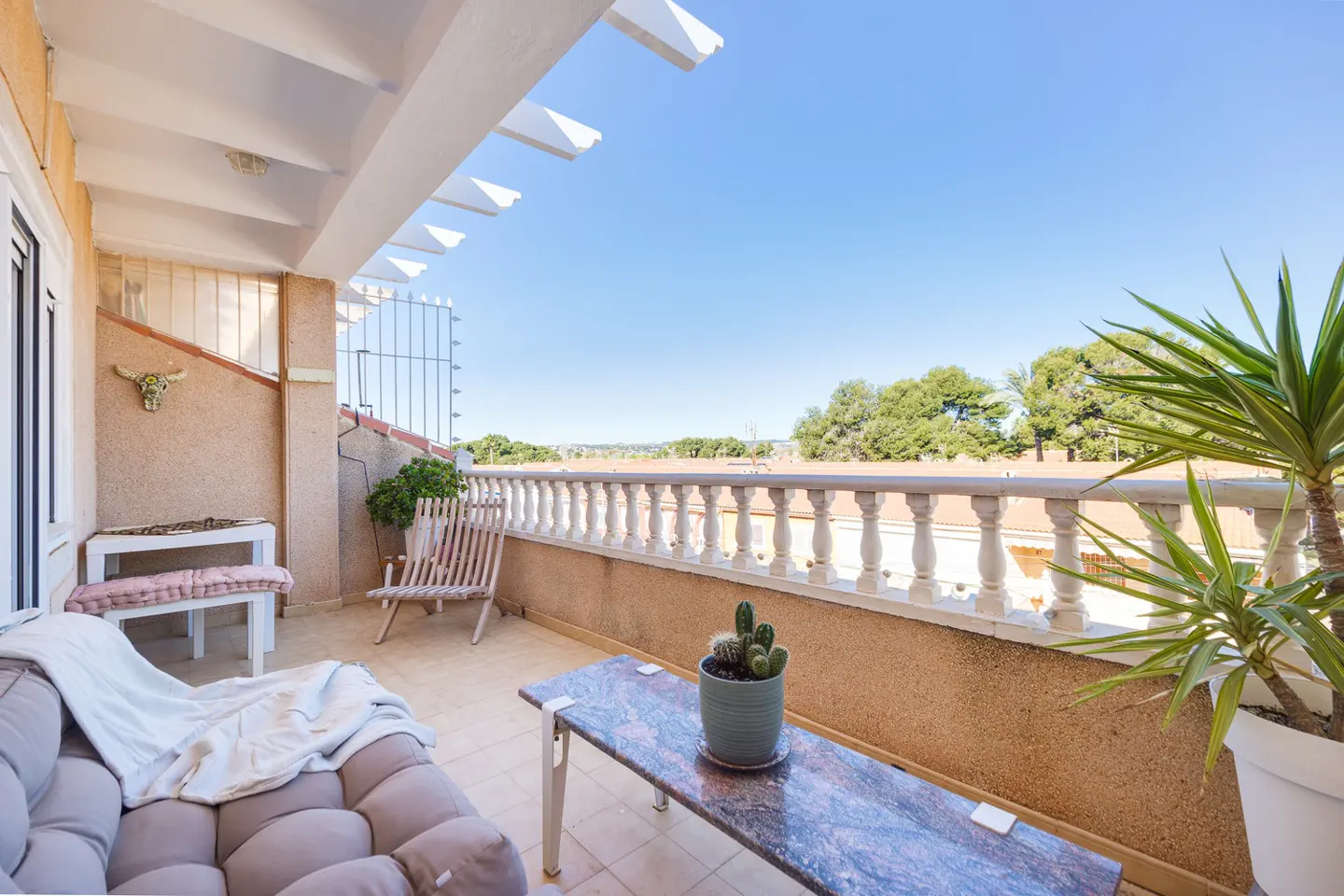 Balcony with a beige sofa, marble table, and white balustrade overlooking trees under a blue sky.