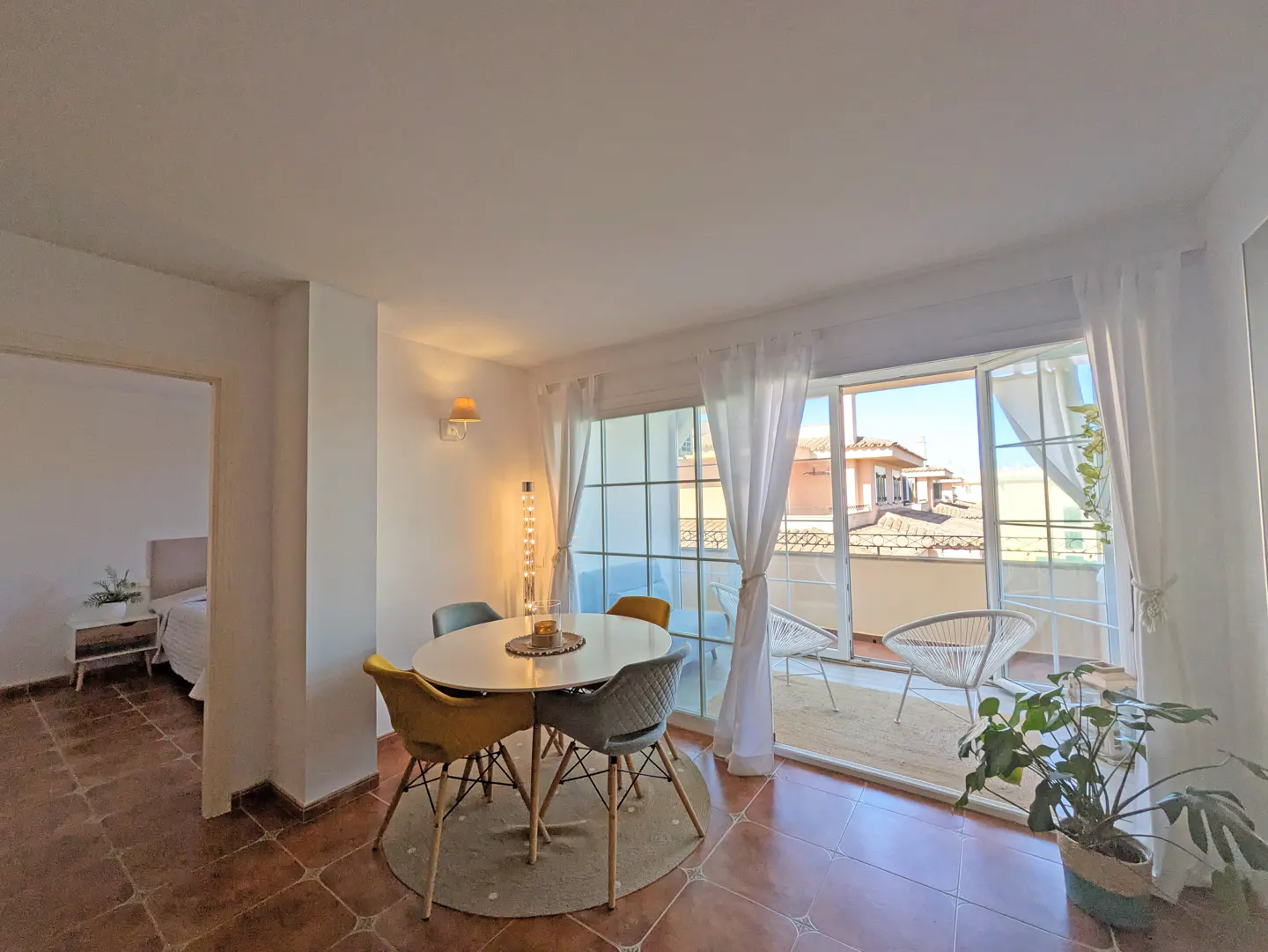 Bright dining area with a round table, colorful chairs, and a balcony with white chairs and a view.