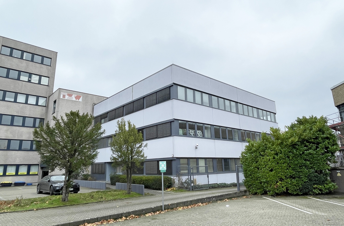 A modern three-story office building with light grey panels, dark window frames, and a parking lot in the foreground. [AI-generated text]