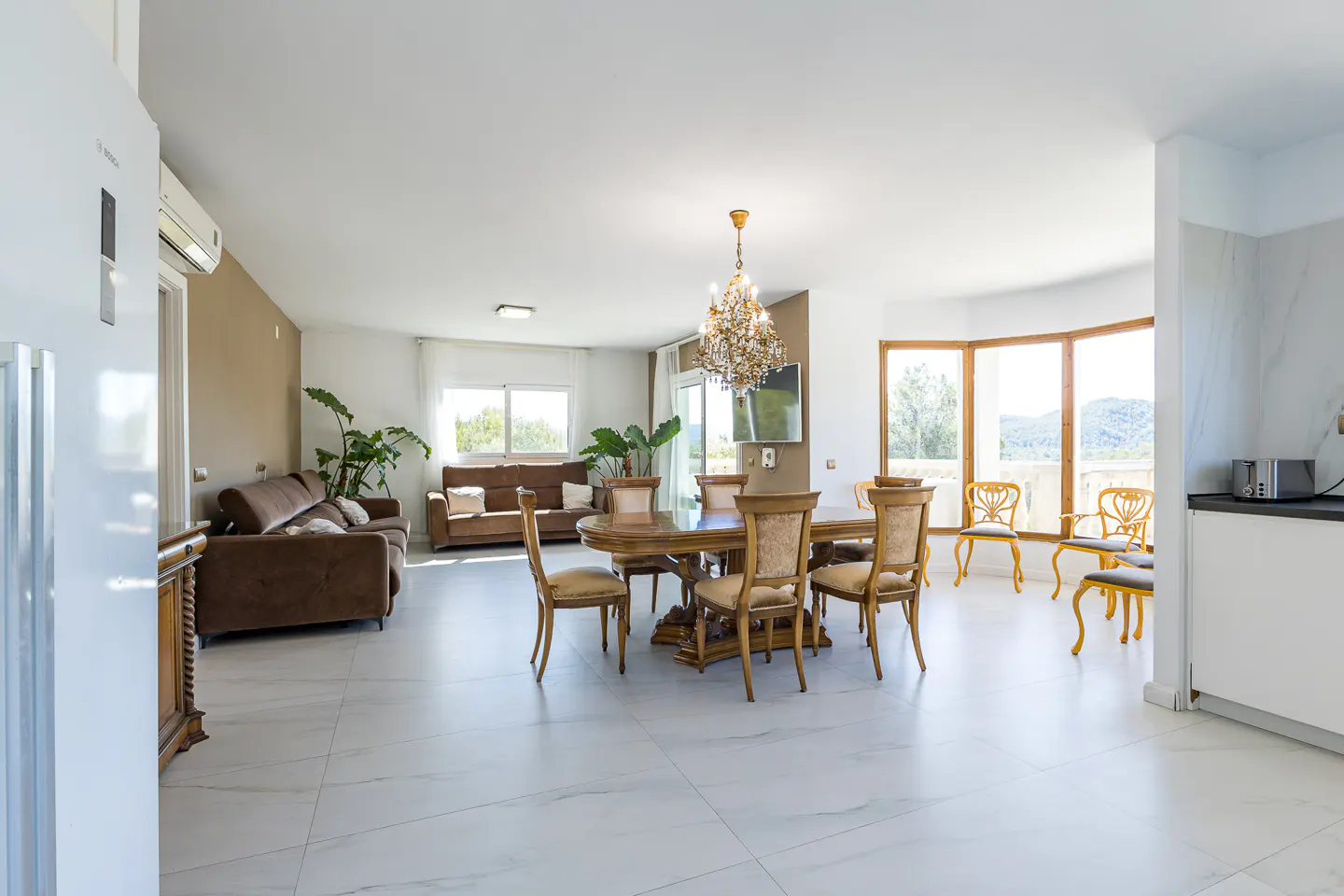 Bright, open-plan living space with marble floors, brown sofas, and a wooden dining table under a chandelier. Large windows offer a scenic view.