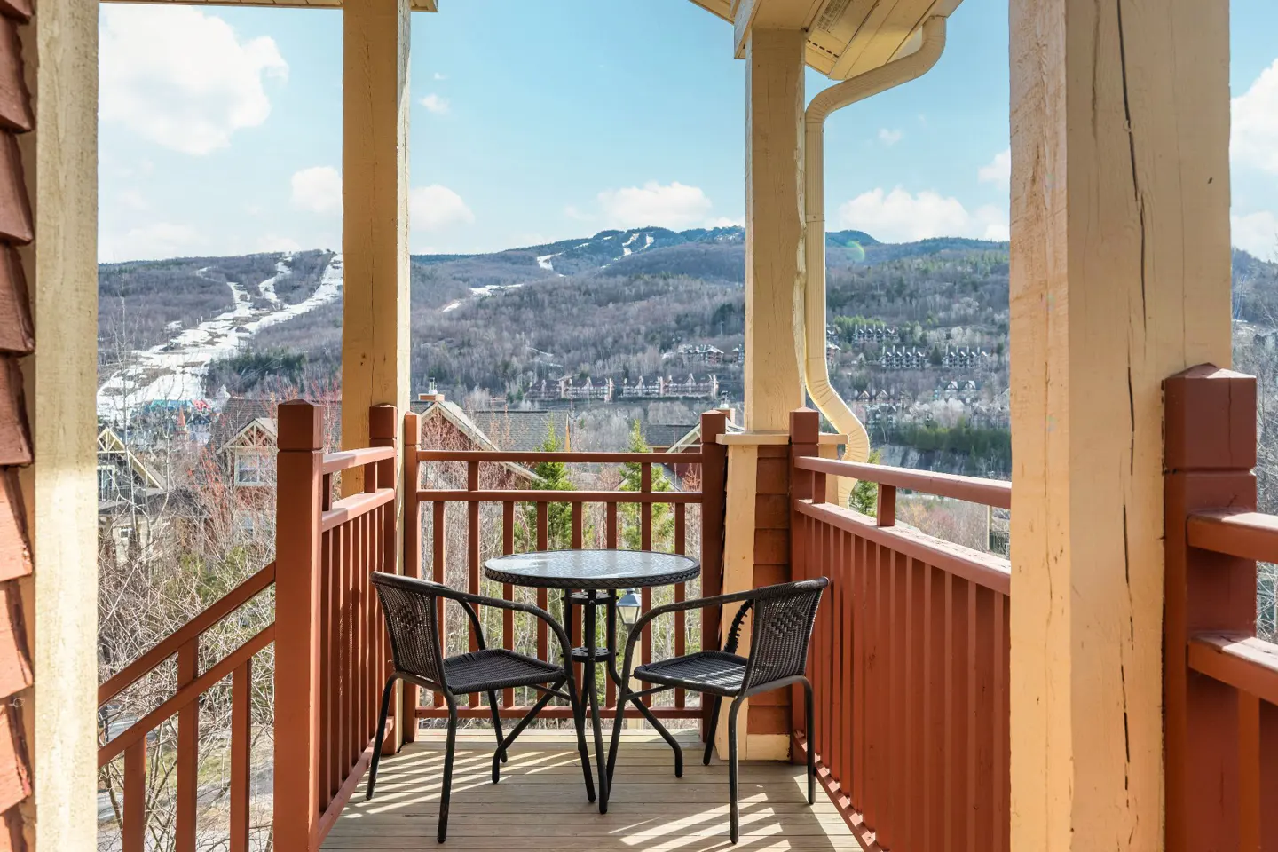 Balcony view with a round table and two chairs overlooking a mountain with ski slopes on a sunny day.