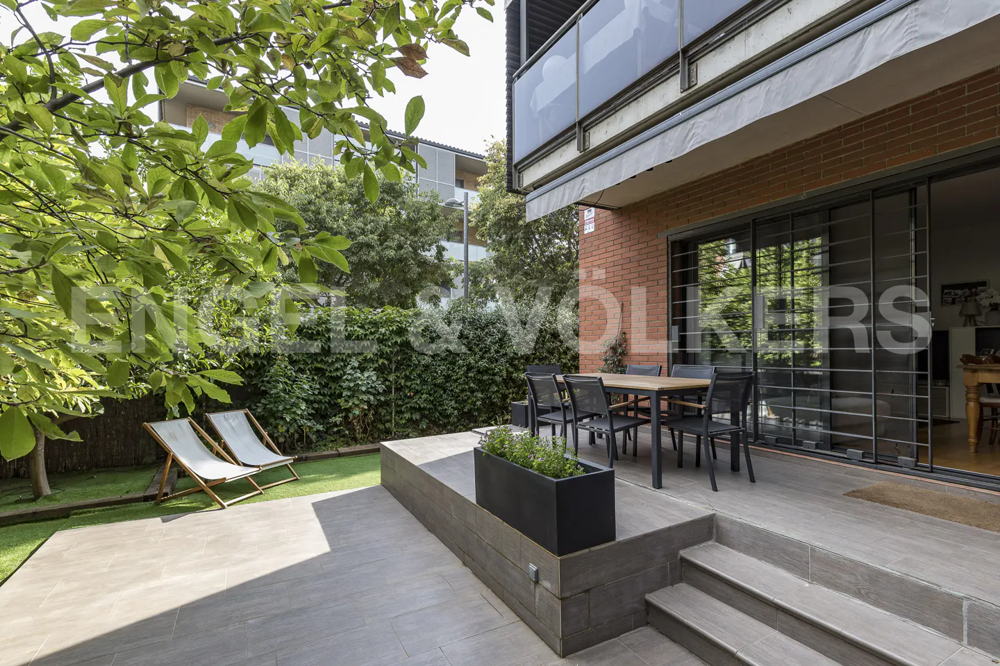 Outdoor patio with a dining table, chairs, and lounge chairs. Brick building with sliding glass doors. Green lawn and trees.