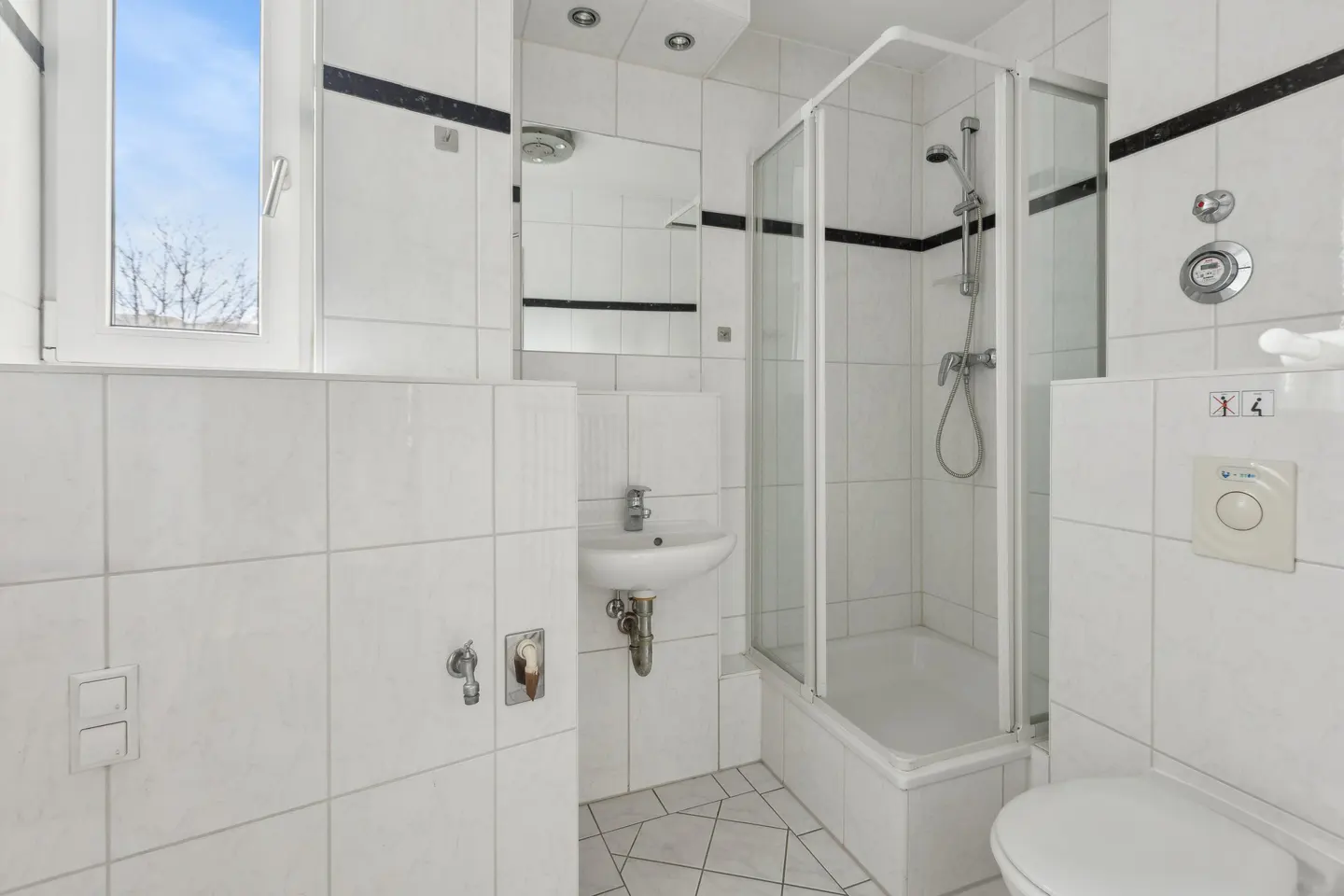 Bright bathroom with white tiles, a glass shower, a sink, and a toilet. A window shows a blue sky.