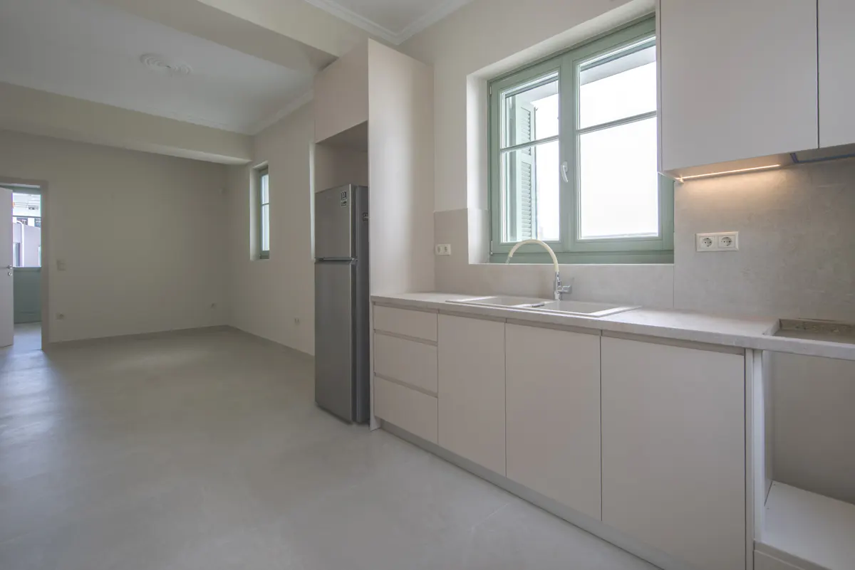 A bright, empty kitchen with white cabinets, a stainless steel refrigerator, and a green-framed window.