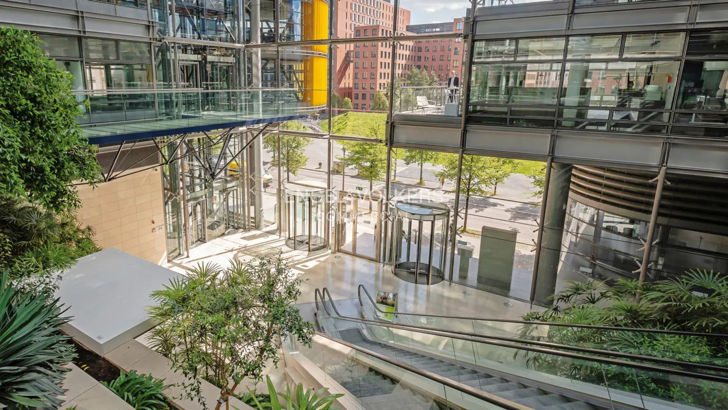 Modern office building interior with glass walls, escalators, revolving doors, and lush green plants.