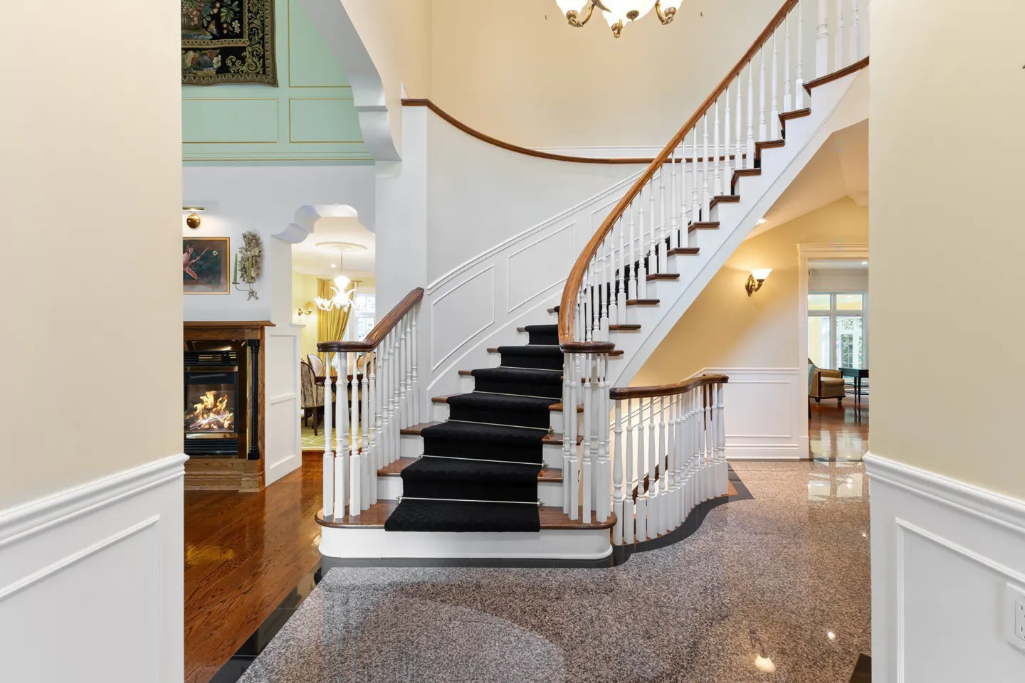 Grand foyer with curved staircase. Black carpeted stairs, white railings, and wood trim. Fireplace visible in adjacent room.