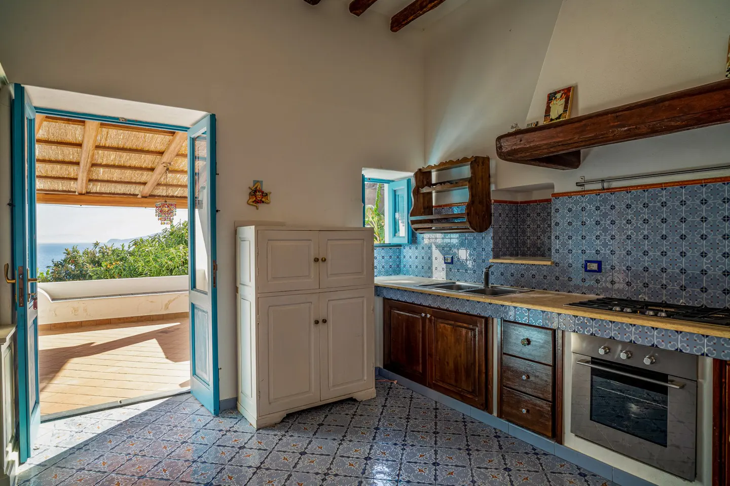 Bright kitchen with blue and white patterned tile floor and backsplash. Open blue door leads to a patio with ocean view.