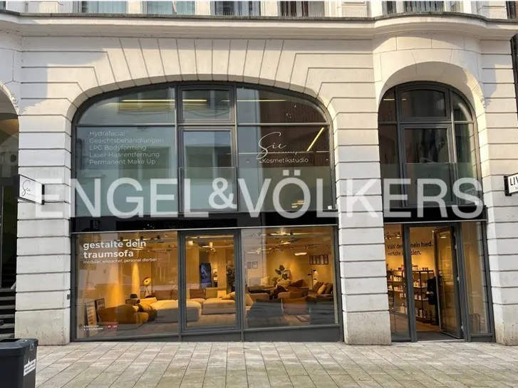Engel & Völkers storefront with large windows displaying furniture and cosmetic services. The building is white stone.