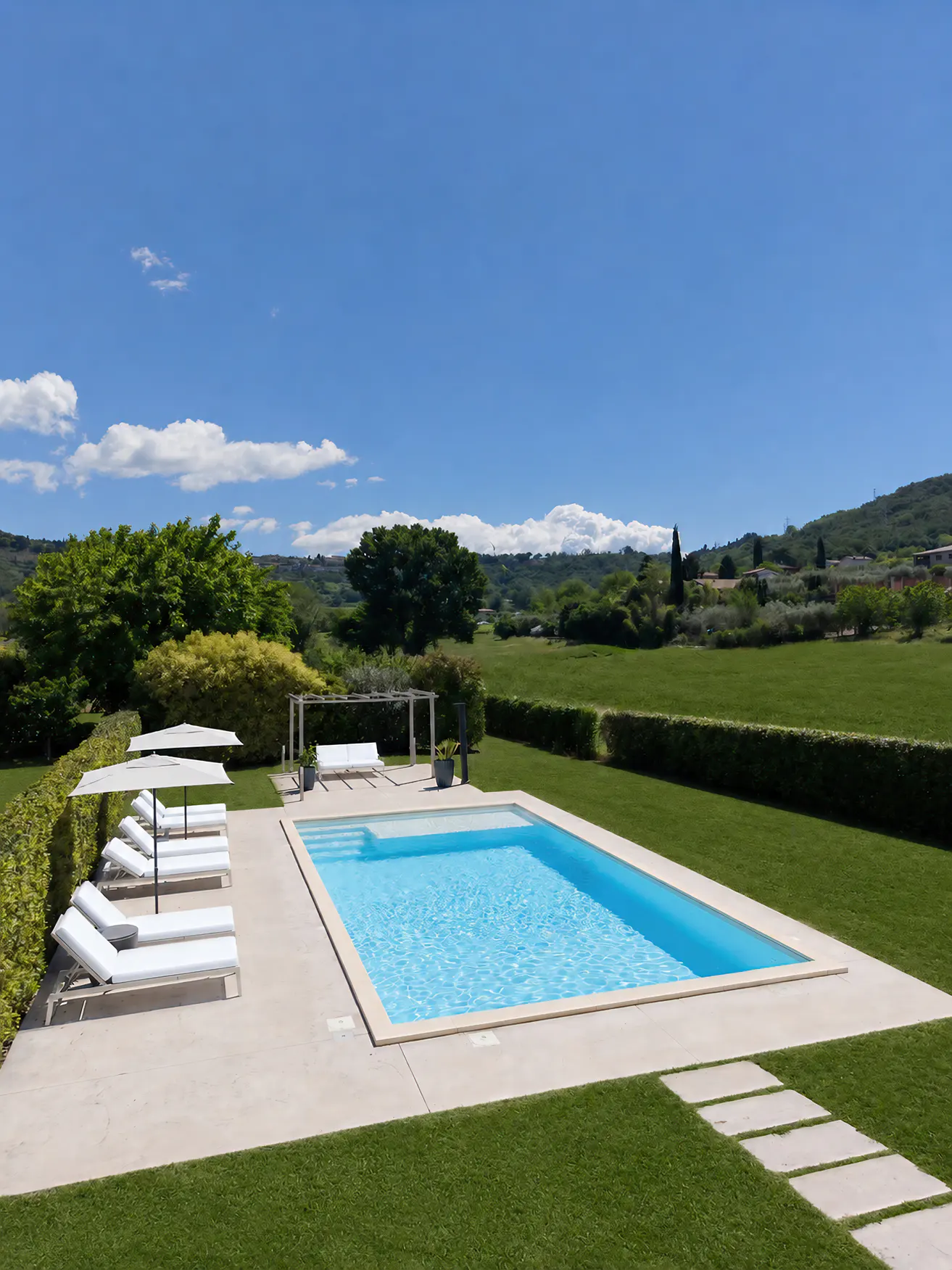 A rectangular swimming pool with white lounge chairs on a patio, surrounded by a green lawn and scenic countryside views.