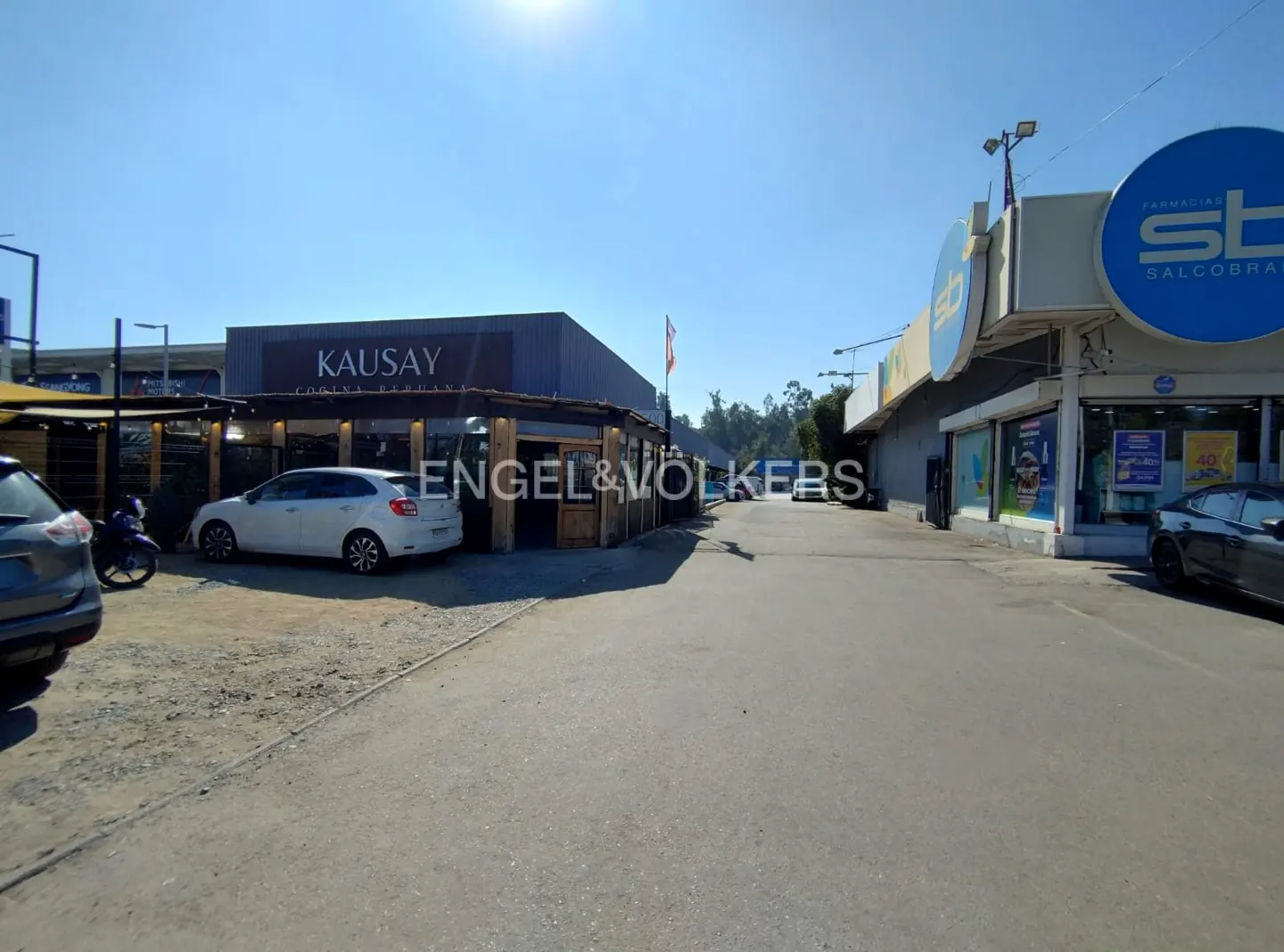 Exterior view of commercial buildings, including a restaurant and pharmacy, with cars parked outside. Engel & Volkers logo visible.
