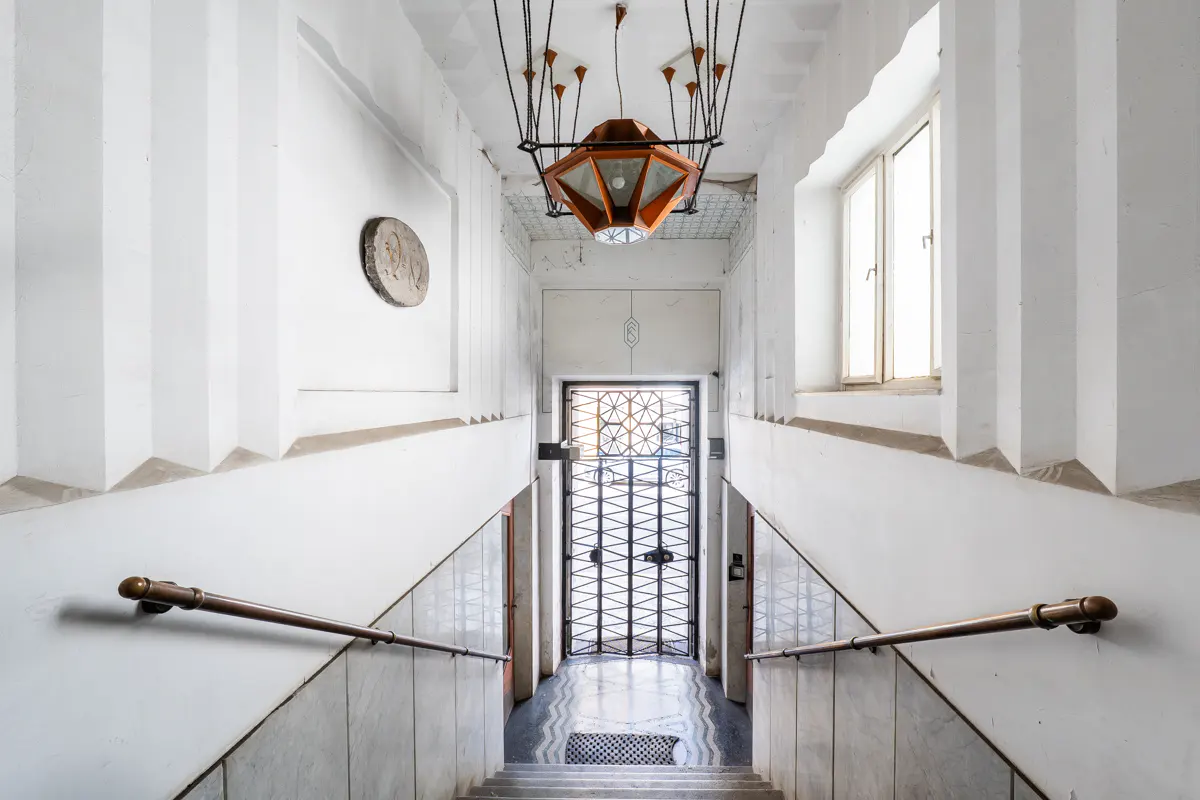 Staircase with white walls, metal handrails, and a geometric pendant light leading to a decorative metal security gate.