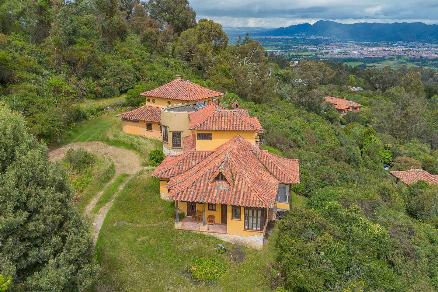 Aerial view of a yellow villa with a red tile roof nestled in a lush, forested hillside overlooking a distant valley.