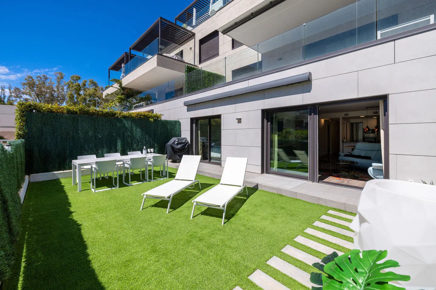 A modern patio with two white lounge chairs on green grass, a dining table, and a grill. The patio is surrounded by a green hedge.