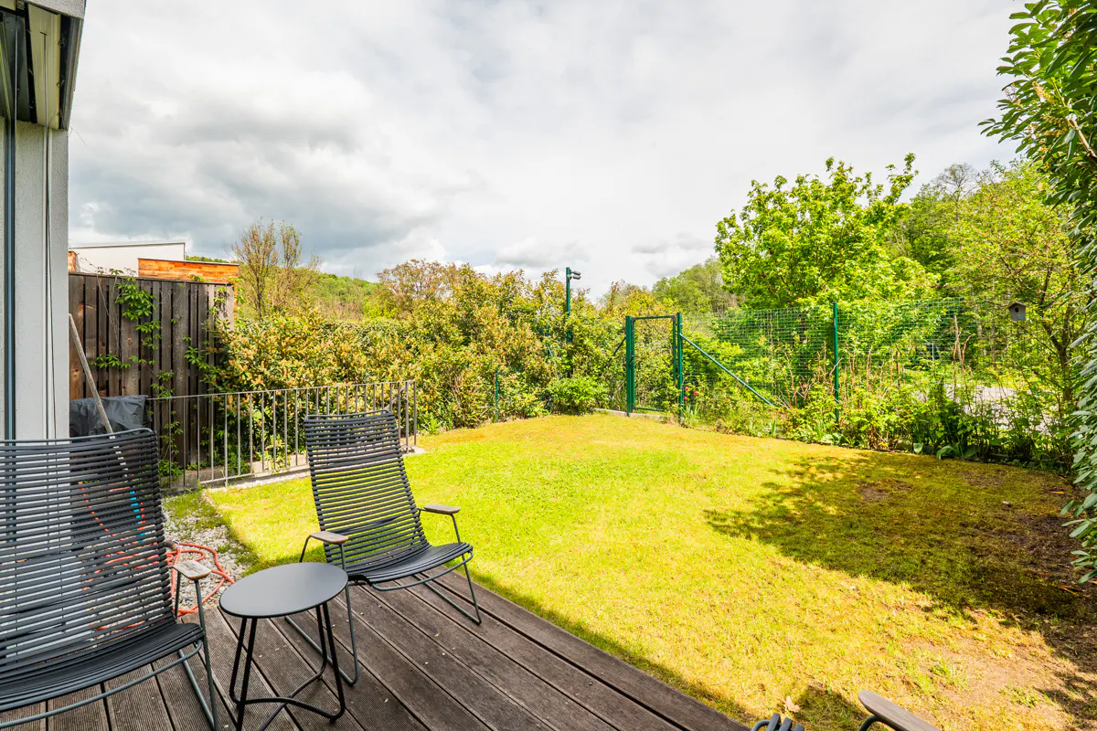 A backyard with a wooden deck, two black chairs, a small table, and a green lawn surrounded by trees and a fence.