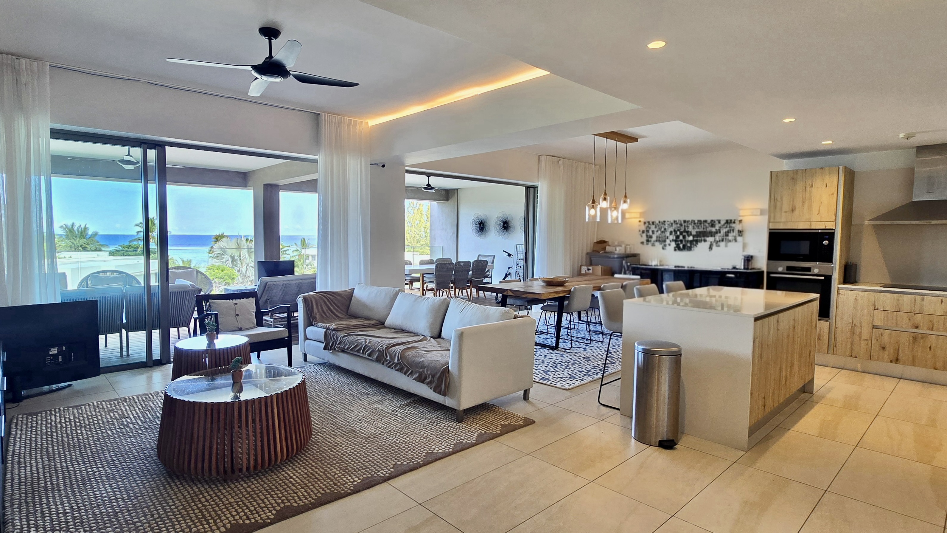 Open-concept living space with a white sofa, dining table, kitchen island, and ocean view through sliding glass doors.