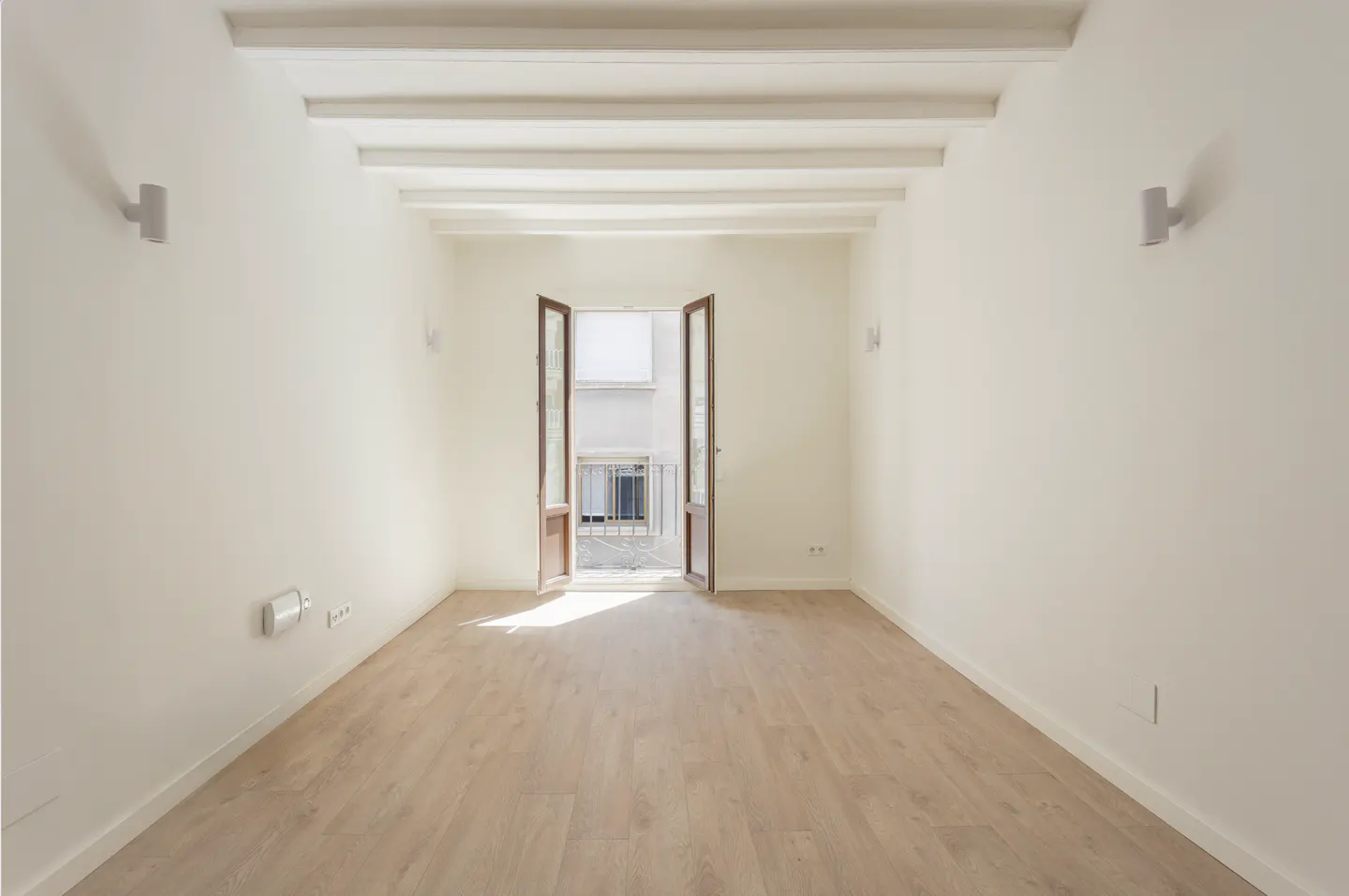 Empty room with white walls, light wood floors, exposed ceiling beams, and open French doors leading to a small balcony.